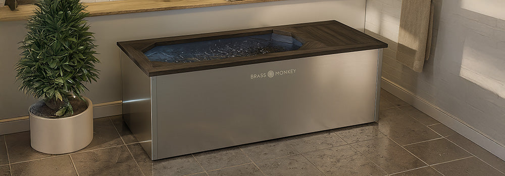 Stainless steel ice bath with dark wood top in modern indoor spa setting with towels and plants.