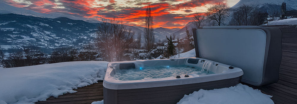 Steaming hot tub on snowy deck with mountain view at sunset.