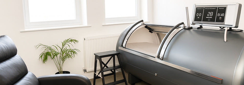 Grey steel hyperbaric oxygen chamber in a bright therapy room with recliner, step stool, and large windows.