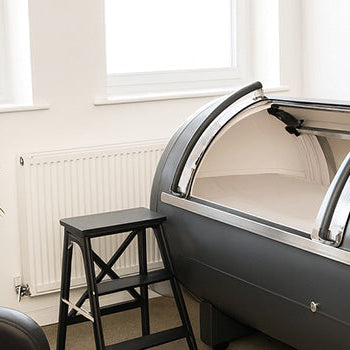 Grey steel hyperbaric oxygen chamber in a bright therapy room with recliner, step stool, and large windows.