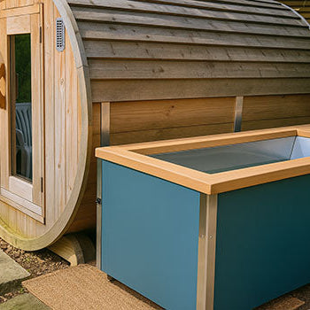 Teal ice bath with wood top beside a round wooden sauna in a private garden with stone path and slatted fencing.