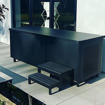 Unwrapped black metal ice bath on a modern tiled patio beside a white contemporary house with glass doors.