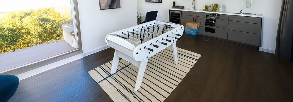 white foosball table on a striped rug in a bright modern room with wall art and a small kitchenette.