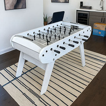 white foosball table on a striped rug in a bright modern room with wall art and a small kitchenette.
