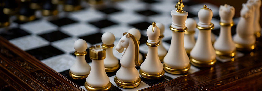 White and gold luxury chess set on ornate carved wood board with black and white tiles in warm lighting.