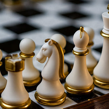White and gold luxury chess set on ornate carved wood board with black and white tiles in warm lighting.