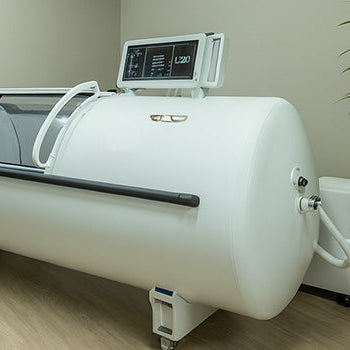 White hyperbaric oxygen chamber in a modern medical treatment room with wood flooring and potted indoor plant.