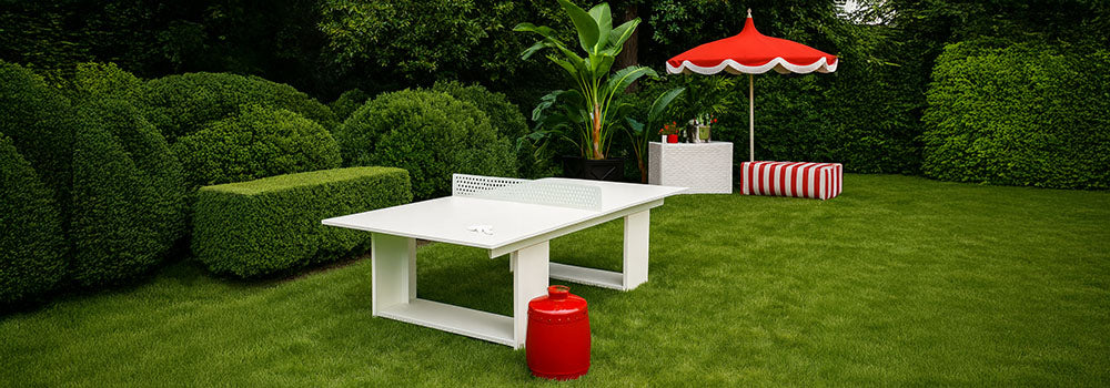 White outdoor table tennis table on lush green lawn with red vintage cooler and striped umbrella in garden setting.