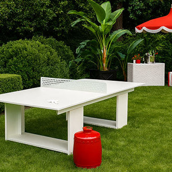 White outdoor table tennis table on lush green lawn with red vintage cooler and striped umbrella in garden setting.