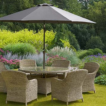 Round wicker dining set with umbrella in a vibrant garden surrounded by colorful flowers and greenery.