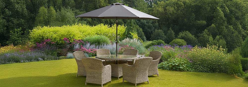Round wicker dining set with umbrella in a vibrant garden surrounded by colorful flowers and greenery.