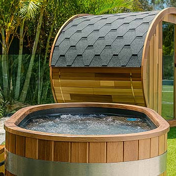 Outdoor wooden ice bath next to barrel sauna surrounded by palm trees and greenery.
