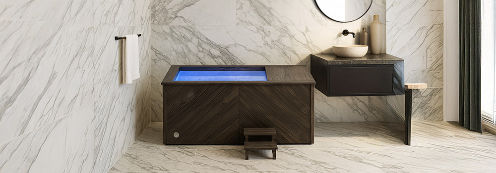 Dark wood ice bath with blue LED lighting in a luxury marble bathroom with stone sink.