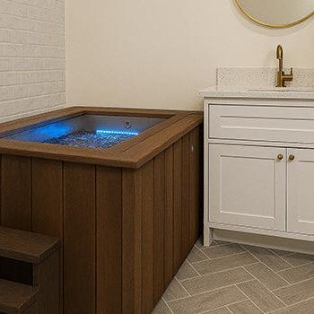 Brown wooden ice bath with blue LED light in modern bathroom with vanity, gold mirror, and herringbone floor.