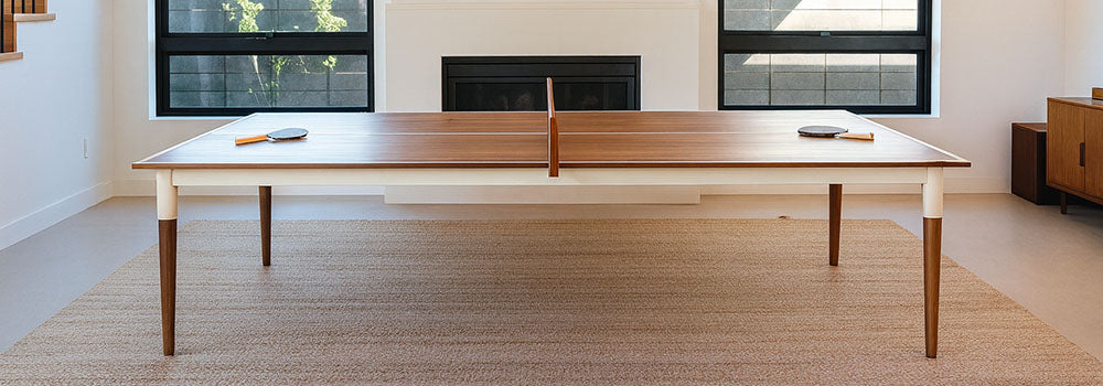 Modern wooden table tennis table with white frame in a bright living room with black-trim windows and fireplace.