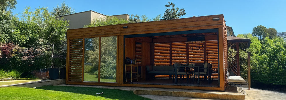 Sleek wooden garden room with open sliding door, modern furniture, and lush backyard setting under blue sky.