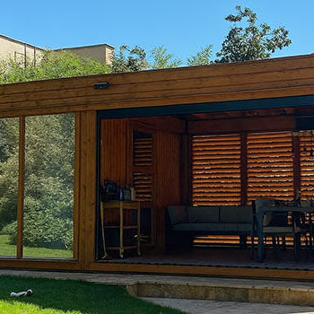 Sleek wooden garden room with open sliding door, modern furniture, and lush backyard setting under blue sky.