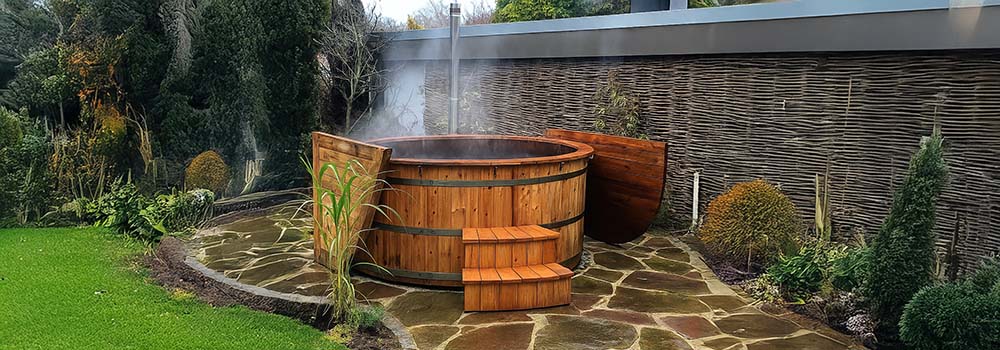 Round wooden hot tub with steam rising on a flagstone patio beside woven fencing and garden greenery.
