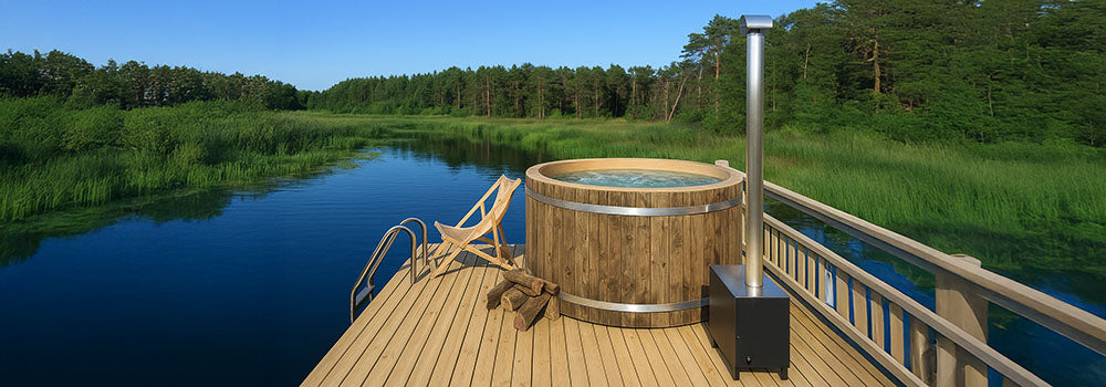 Rustic wooden hot tub on a lakeside deck with firewood and chair under clear blue sky.