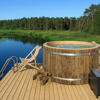 Rustic wooden hot tub on a lakeside deck with firewood and chair under clear blue sky.