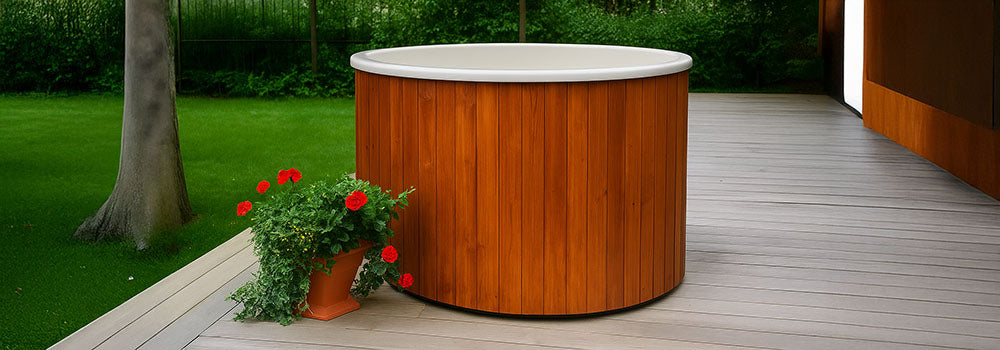 Round wooden hot tub on outdoor deck with flower planter and green garden background.