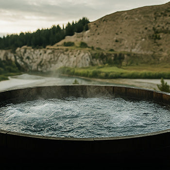 Round wooden hot tub steaming on a deck with a panoramic view of a river winding through a mountain valley.