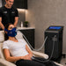 Female client wearing an Altitude Centre MetraFit Pro breathing mask during a live hypoxic training session, supervised by a male trainer, with the aetraFit touchscreen display showing real-time oxygen saturation and session progress