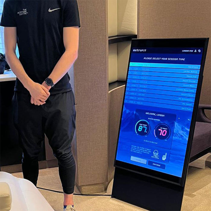 Altitude Centre trainer standing beside the MetraFit Pro vertical touchscreen display showing the session type selection interface with altitude-based training levels