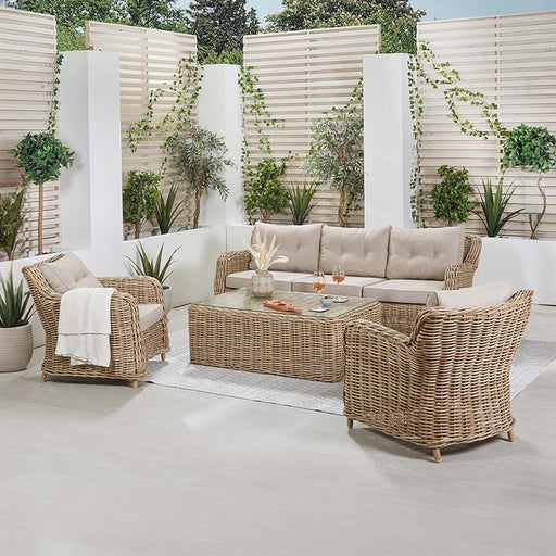 Barakaldo Natural Antique Outdoor Seating Set arranged in a garden-inspired indoor space, featuring a rattan sofa, armchairs, and a glass-top coffee table with beige cushions.