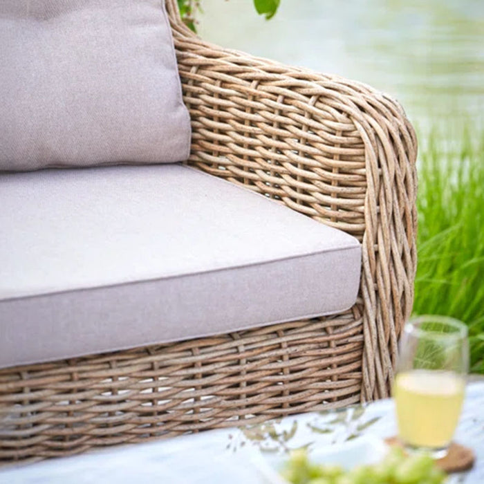 Close-up of the Barakaldo Natural Antique Outdoor Seating Set's sofa arm, highlighting the intricate rattan weave and beige cushion, set against a peaceful outdoor backdrop.