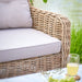 Close-up of the Barakaldo Natural Antique Outdoor Seating Set's sofa arm, highlighting the intricate rattan weave and beige cushion, set against a peaceful outdoor backdrop.