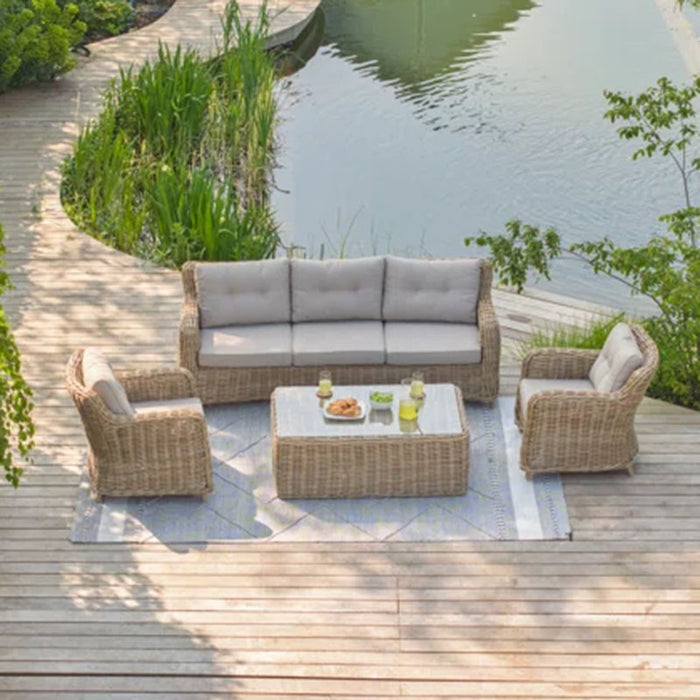 Barakaldo Natural Antique Outdoor Seating Set placed on a lakeside deck, offering a serene view with a rattan sofa, armchairs, and a coffee table.