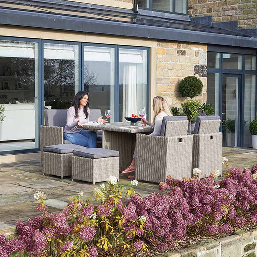 Two women enjoying drinks and conversation at the Stone Grey Bermuda Cube Set with Ceramic Top in a garden setting.