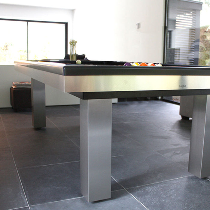 Polished stainless steel frame pool table with black cloth in a minimalistic tiled living area with natural light.