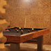Warm-toned Billards Toulet Megeve Modern pool table with black felt in a stone-walled room featuring stacked wood decor and ambient lighting.