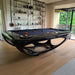 Glossy black Billards Toulet Whitelight pool table featured in a cosy, wood-floored room with a view of greenery and stylish shelf decor for a serene vibe.