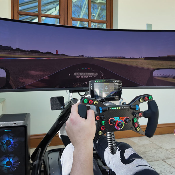 Driver's point of view on a BPS Blade Racing Simulator showing hands gripping a carbon fibre racing steering wheel with ultrawide curved monitor displaying a circuit