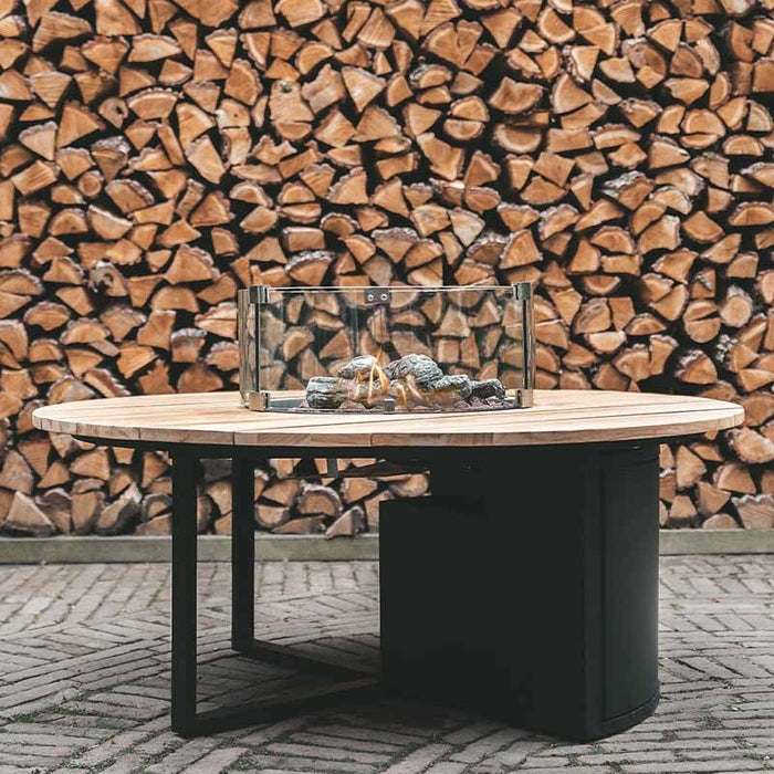 Cosiloft 120 Round Fire Pit Table set up in a stylish backyard with a stack of firewood in the background, showcasing the elegant combination of a black aluminium frame and teak wood top.