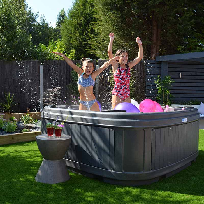 Kids relaxes in a dark grey hot tub set on a lawn, with a blue lit water feature.