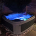 Dark grey hot tub installed on a brick patio, lit up at night with blue lights.