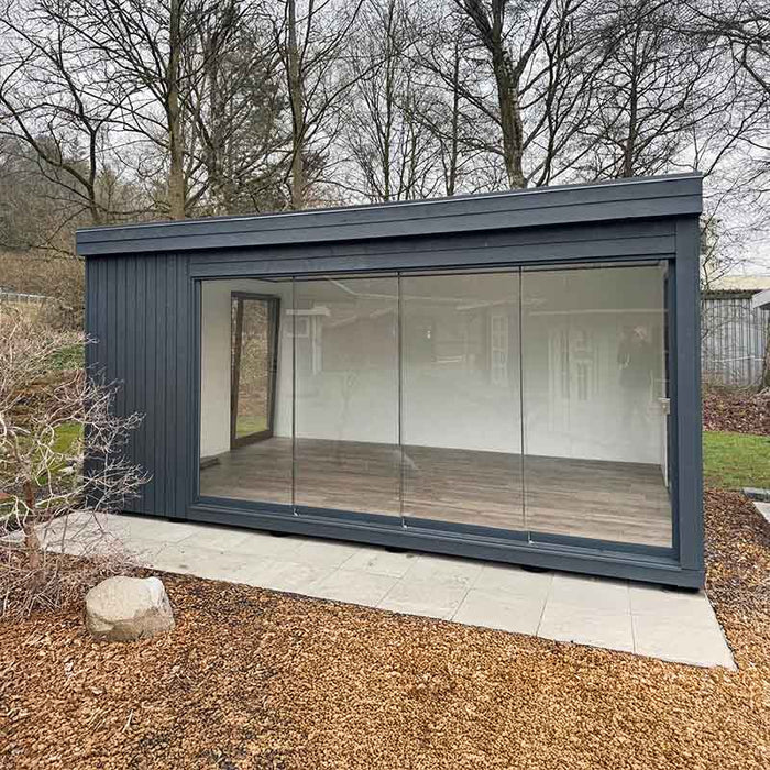 Dark-painted Estholz Garden House WILLIAM with a full-length glass wall, positioned in a quiet, leafless garden landscape.