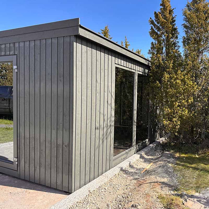 Side view of Estholz Garden House WILLIAM with grey vertical timber cladding and tall glass windows blending into a wooded area.