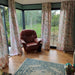 Cosy interior of Estholz Summerhouse WALTER featuring floral curtains, large panoramic windows, and a leather armchair overlooking the peaceful garden setting.