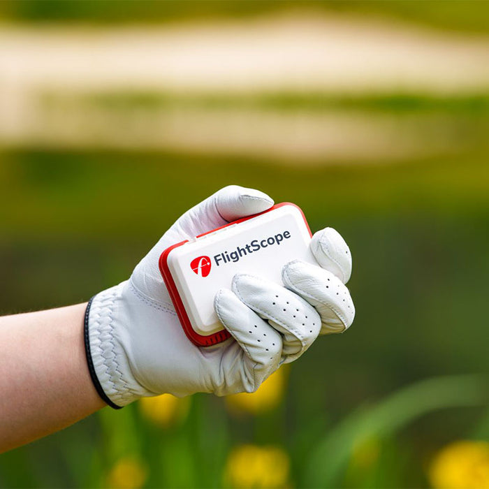 Hand holding FlightScope Mevo launch monitor against a grass backdrop.