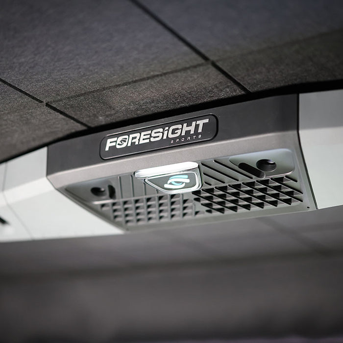 Front close-up of Foresight Falcon overhead launch monitor showing glowing tracking sensor and vent grid, mounted on a black panel ceiling.