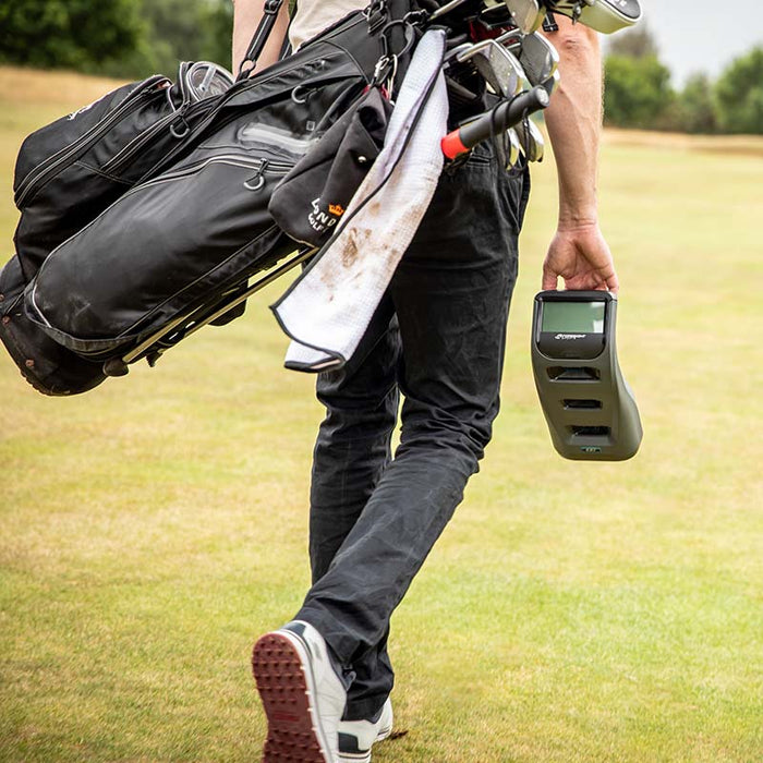Golfer walking across the fairway carrying the Foresight GC3 Launch Monitor by hand alongside a golf bag.
