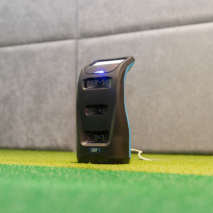 Indoor simulator setup featuring the Foresight GC3S Launch Monitor with blue indicator light activated on padded wall background.