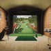 Interior view of Foresight Sim-in-a-Box Play Golf Simulator in brick-walled space, featuring dual golf bags, launch monitor, and immersive course graphics.