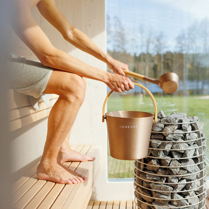 Person pouring water over hot sauna stones using a Geneva-branded ladle and bucket, inside the St. Moritz outdoor sauna with panoramic view.