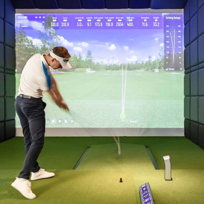 A golfer taking a swing indoors with the Golfjoy GDS Plus Launch Monitor, displaying detailed shot data on the screen.